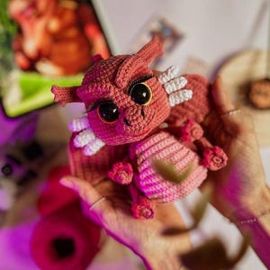 May include: A handmade crochet dragon in shades of pink and red. The dragon has large black eyes, white accents, and small horns. The dragon is held in someone's hands.