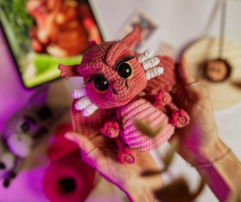May include: A handmade crochet dragon in shades of pink and red. The dragon has large black eyes, white accents, and small horns. The dragon is held in someone's hands.