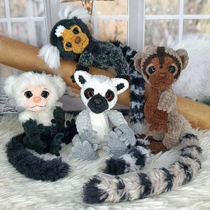 May include: A collection of handmade plush lemurs and monkeys in various colors, including white, gray, black, and brown. The plush toys have detailed features and long tails, displayed on a white, fluffy surface.