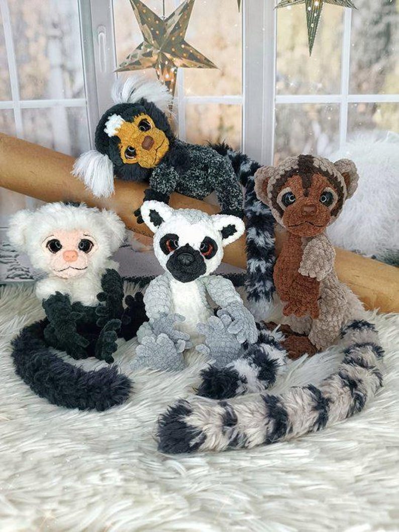 May include: A collection of handmade plush lemurs and monkeys in various colors, including white, gray, black, and brown. The plush toys have detailed features and long tails, displayed on a white, fluffy surface.