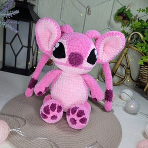 May include: A pink plush toy with large ears, black eyes, and purple accents on the face, feet, and arms. The stuffed animal is made of a soft, textured material and is sitting on a neutral-colored surface. The toy is a character from a popular animated film.