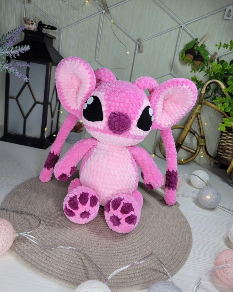 May include: A pink plush toy with large ears, black eyes, and purple accents on the face, feet, and arms. The stuffed animal is made of a soft, textured material and is sitting on a neutral-colored surface. The toy is a character from a popular animated film.