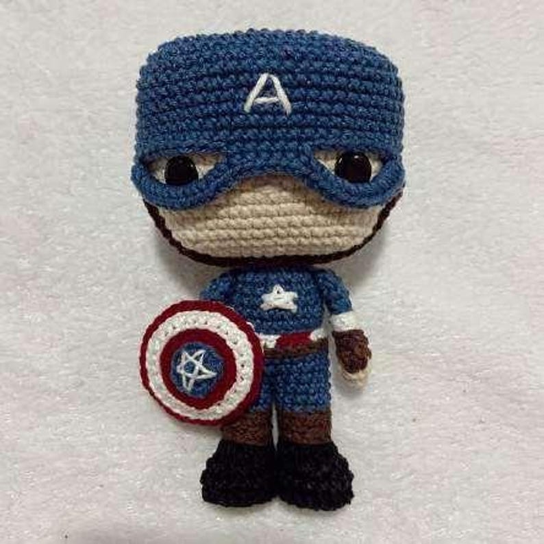 May include: A handmade Captain America amigurumi doll. The doll is crocheted with blue, beige, red, white, brown, and black yarn. It features a blue helmet with a white "A", a shield, and a star on the chest.