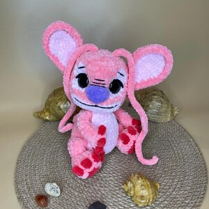 May include: A handmade pink plush toy with large ears, black eyes, and a purple nose. The plushie has red spots on its feet and is sitting on a round, woven mat. Decorative seashells and small stones surround the toy.