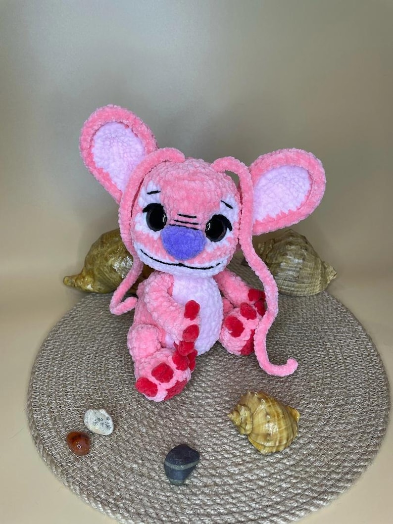 May include: A handmade pink plush toy with large ears, black eyes, and a purple nose. The plushie has red spots on its feet and is sitting on a round, woven mat. Decorative seashells and small stones surround the toy.