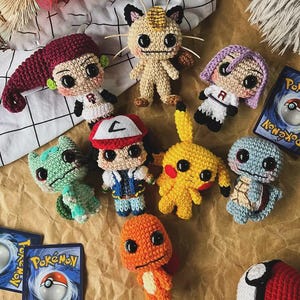 May include: A collection of handmade crochet Pok&eacute;mon characters, including Pikachu, Charmander, and Squirtle, along with two Pok&eacute;mon trading cards and a Pok&eacute; Ball. The figures are in various colors and sizes, arranged on a textured surface.