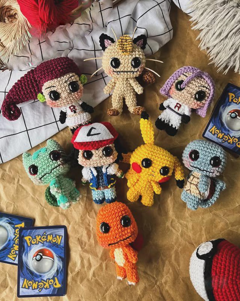 May include: A collection of handmade crochet Pok&eacute;mon characters, including Pikachu, Charmander, and Squirtle, along with two Pok&eacute;mon trading cards and a Pok&eacute; Ball. The figures are in various colors and sizes, arranged on a textured surface.