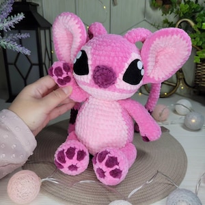 May include: A handmade pink plush toy with large ears, black eyes, and a purple nose and feet. The toy is made of soft, textured yarn and has a friendly, cartoonish appearance. The toy is a character from a popular animated film.