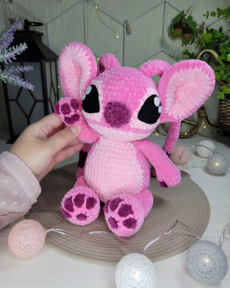 May include: A handmade pink plush toy with large ears, black eyes, and a purple nose and feet. The toy is made of soft, textured yarn and has a friendly, cartoonish appearance. The toy is a character from a popular animated film.