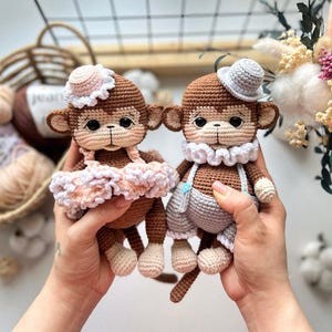 Peut inclure: Deux poupées singes crochetées à la main. L'une porte un tutu rose et blanc et un chapeau assorti. L'autre porte une salopette grise et un chapeau blanc. Les deux poupées ont un corps marron, un visage beige et des yeux noirs.
