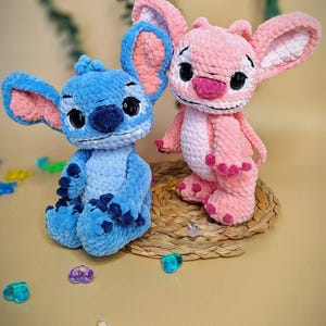 May include: Two handmade crochet plush toys, one blue and one pink, resembling a cartoon character. The blue toy has blue and pink ears, and the pink toy has pink and white ears. Both have black eyes and are sitting on a woven mat.