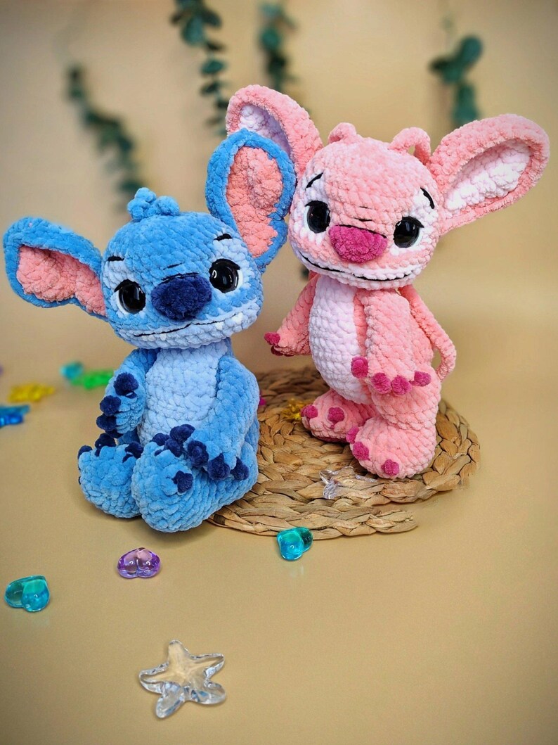 May include: Two handmade crochet plush toys, one blue and one pink, resembling a cartoon character. The blue toy has blue and pink ears, and the pink toy has pink and white ears. Both have black eyes and are sitting on a woven mat.