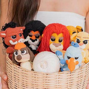 Könnte beinhalten: Ein Weidenkorb gefüllt mit handgefertigten gehäkelten Tierpuppen. Die Puppen umfassen einen Löwen mit roter Mähne, einen Hund mit orangefarbener Mütze und einen blauen Vogel. Ein Knäuel weißer Wolle liegt im Korb.