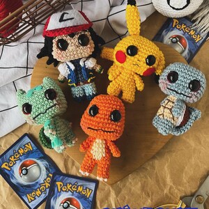 May include: A collection of handmade crochet Pok&eacute;mon characters, including Pikachu, Bulbasaur, Charmander, Squirtle, and Ash Ketchum. The characters are in various colors and sizes, with large black eyes. The image also includes Pok&eacute;mon trading cards and a Pok&eacute; Ball.