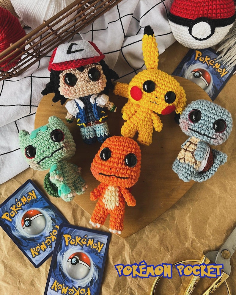 May include: A collection of handmade crochet Pok&eacute;mon characters, including Pikachu, Bulbasaur, Charmander, Squirtle, and Ash Ketchum. The characters are in various colors and sizes, with large black eyes. The image also includes Pok&eacute;mon trading cards and a Pok&eacute; Ball.
