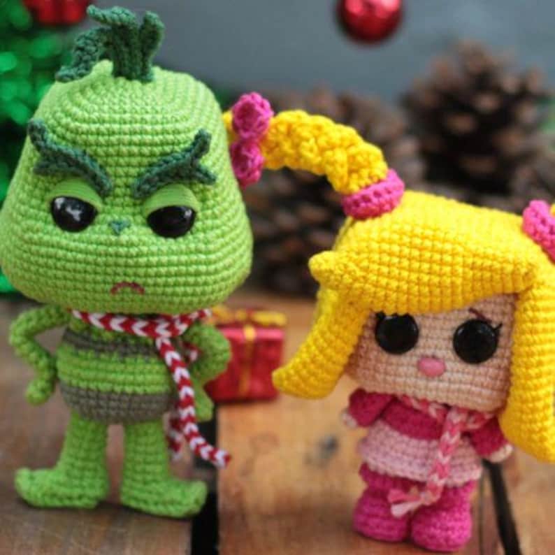 May include: Two crocheted dolls are featured in this image. One is green with a red and white scarf, and the other is yellow and pink. The dolls have black eyes and are standing on a wooden surface. A small red gift box is in the background.