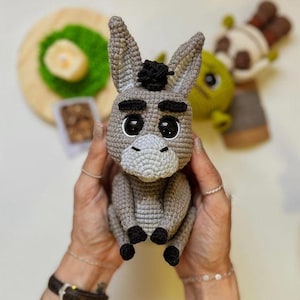 May include: A handmade crochet donkey doll in shades of gray, with black accents on the hooves and a small black tuft of hair. The donkey has large, expressive eyes and a light gray muzzle. Other crochet dolls are visible in the background.