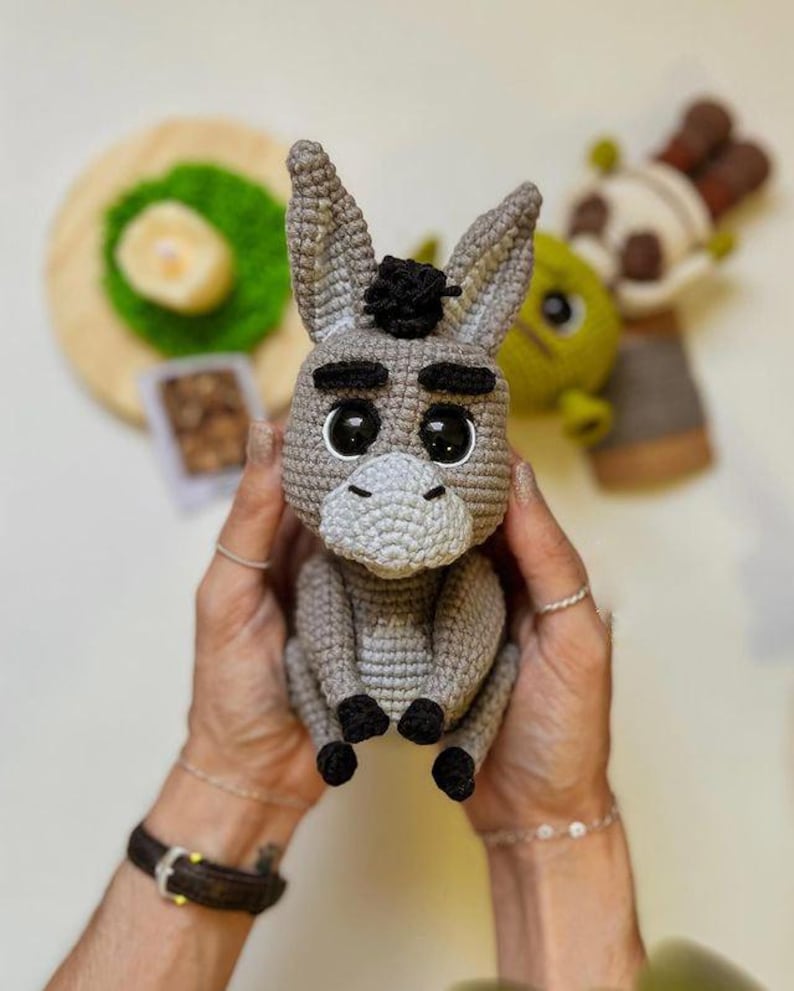 May include: A handmade crochet donkey doll in shades of gray, with black accents on the hooves and a small black tuft of hair. The donkey has large, expressive eyes and a light gray muzzle. Other crochet dolls are visible in the background.