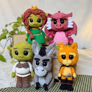 Peut inclure: Un ensemble de cinq poupées en crochet faites à la main comprenant un ogre vert, une ogresse aux cheveux roux, un dragon rose, un âne gris et un chat orange. Chaque personnage a de grands yeux noirs et des détails en laine cousus main.