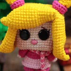 May include: A handmade crochet doll with yellow hair styled in pigtails, accented with pink bands. The doll has large black button eyes, a pink nose, and is wearing a pink and white striped dress and scarf. The doll is a charming handmade item.