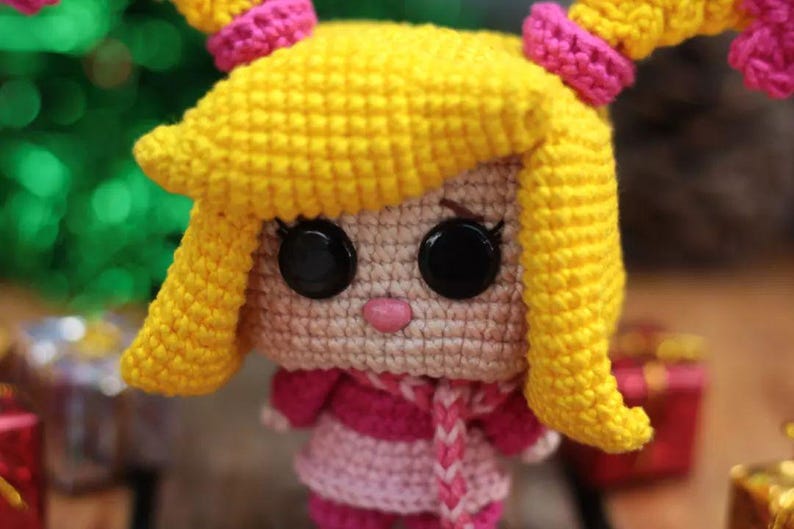 May include: A handmade crochet doll with yellow hair styled in pigtails, accented with pink bands. The doll has large black button eyes, a pink nose, and is wearing a pink and white striped dress and scarf. The doll is a charming handmade item.