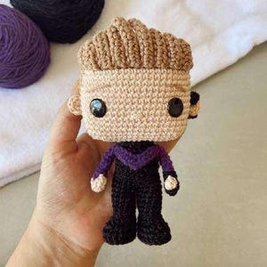 May include: A handmade crochet doll with a light tan head and brown hair. The doll has black eyes, a purple and black outfit, and is being held in a hand. The doll's features are carefully crafted, showcasing the artistry of crochet.