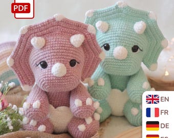 May include: Two crocheted triceratops toys, one in dusty rose and one in mint green, both featuring white horns, white claws, and dark safety eyes. A PDF icon is visible in the top left corner, and a language selection menu is in the bottom right.