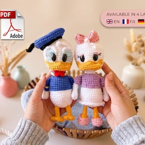 Könnte beinhalten: Zwei gehäkelte Puppen, die eine Entenfigur in einem blauen Matrosenoutfit mit passender Mütze und eine Entenfigur in einem lila Oberteil und weißem Rock mit rosa Schleife darstellen. Das Bild enthält ein PDF-Symbol und Text, der in 4 Sprachen verfügbar ist.
