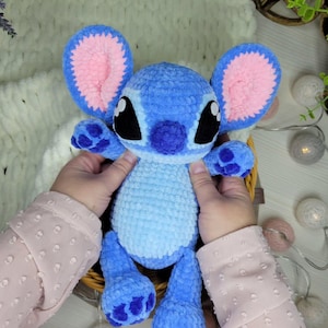 May include: A handmade blue plush toy with large ears, black eyes, and a pink inner ear design. The plush has a light blue body and darker blue accents on the feet and hands. The toy is held in a wicker basket.
