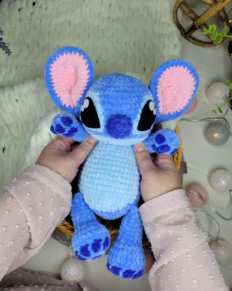 May include: A handmade blue plush toy with large ears, black eyes, and a pink inner ear design. The plush has a light blue body and darker blue accents on the feet and hands. The toy is held in a wicker basket.