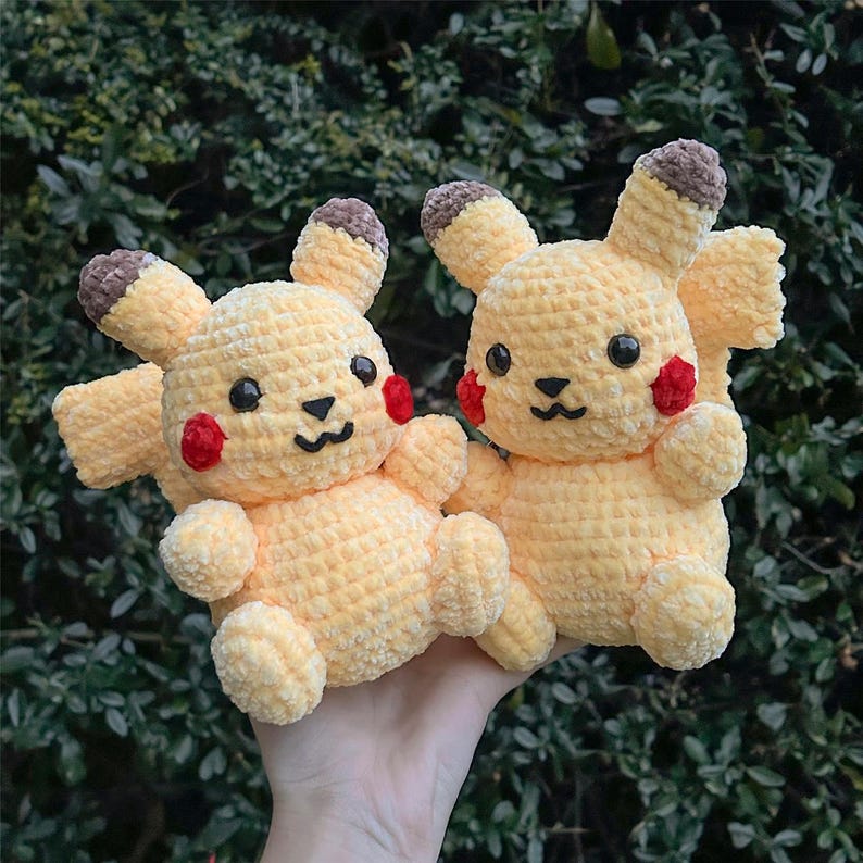 May include: Two crocheted yellow Pikachu plushies with red cheeks and black eyes. The plushies are sitting on a hand.
