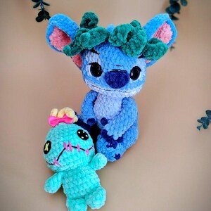 May include: Two handmade crochet plush toys. One is blue with a green headpiece and pink inner ears. The other is turquoise with pink stitching and a yellow bow. Both have black button eyes.
