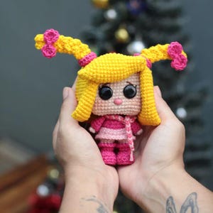 May include: A handmade crochet doll with yellow hair styled in two pigtails, each adorned with pink bows. The doll wears a pink and white striped dress, a pink scarf, and pink boots. The doll has large black eyes and a pink nose.