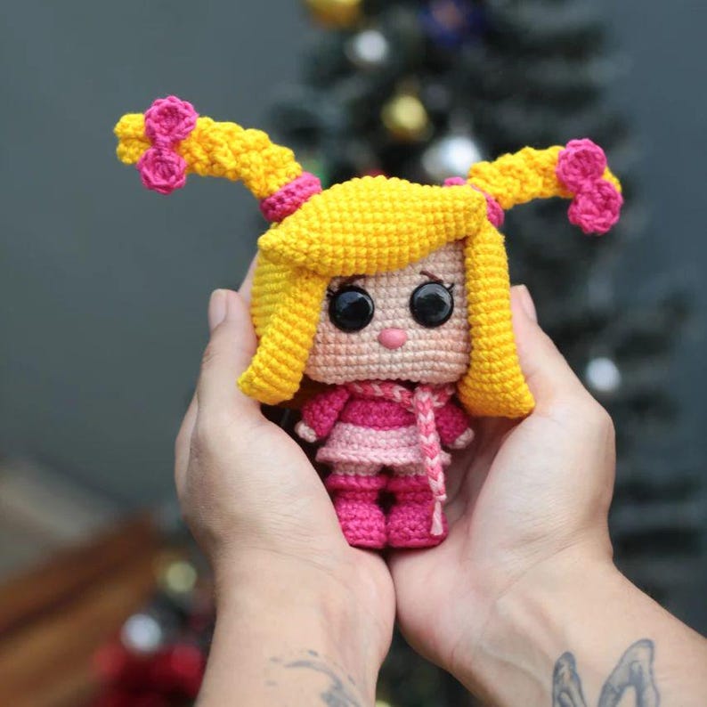 May include: A handmade crochet doll with yellow hair styled in two pigtails, each adorned with pink bows. The doll wears a pink and white striped dress, a pink scarf, and pink boots. The doll has large black eyes and a pink nose.