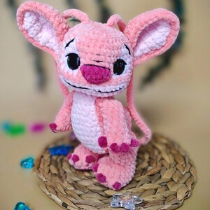 May include: A handmade pink plush toy of a cartoon character with large ears, black eyes, and a dark pink nose. The plush is sitting on a woven mat with colorful decorative gems scattered around it.