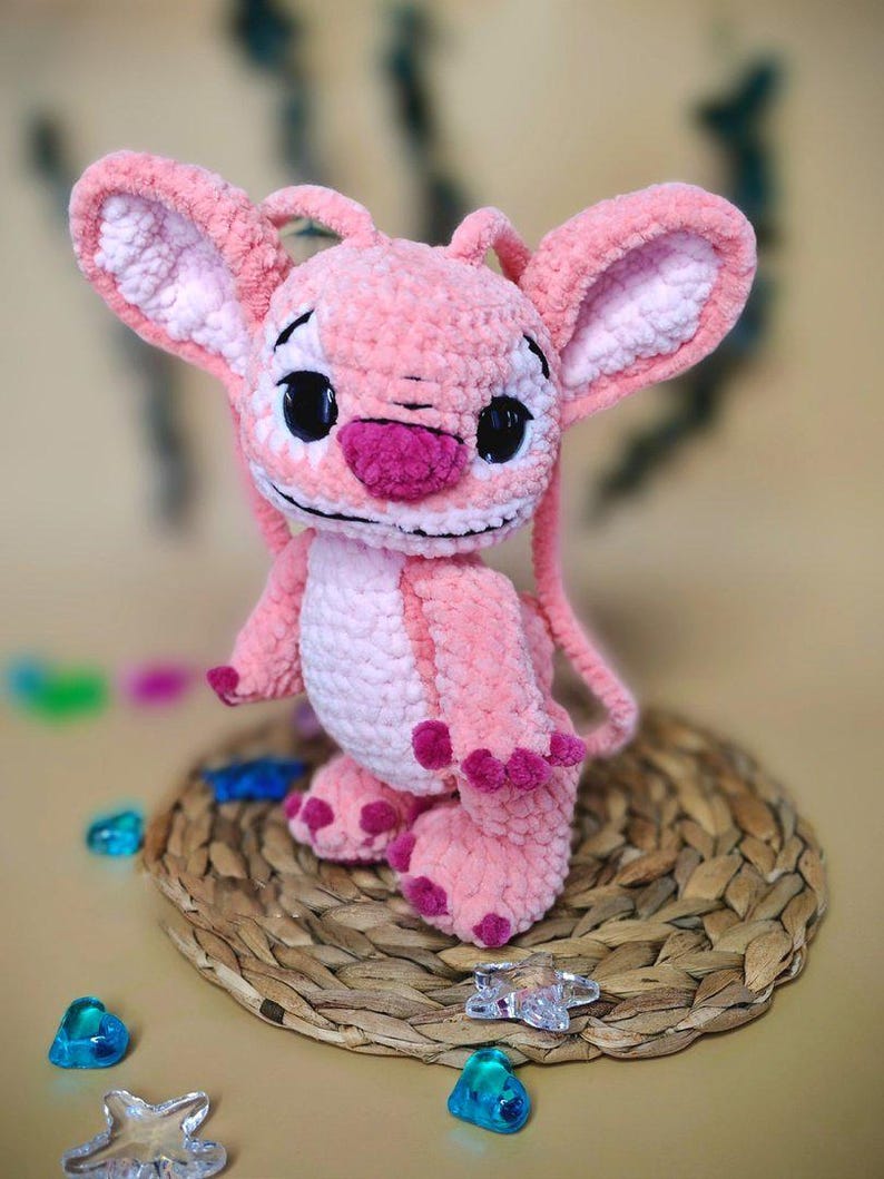 May include: A handmade pink plush toy of a cartoon character with large ears, black eyes, and a dark pink nose. The plush is sitting on a woven mat with colorful decorative gems scattered around it.
