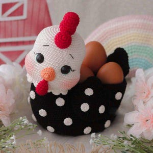 Peut inclure: Un panier à œufs en forme de poule crocheté à la main. La poule a une tête blanche avec une crête et un barbillon rouges, et un corps noir à pois blancs. Le panier est rempli d'œufs bruns. L'arrière-plan comprend une grange rose et un arc-en-ciel.
