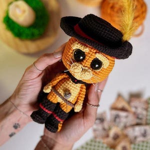 May include: A handmade crochet cat doll, featuring an orange body, black boots, and a black hat with a red band and yellow feather. The doll has large black eyes and white whiskers. The cat is wearing a belt with a buckle.