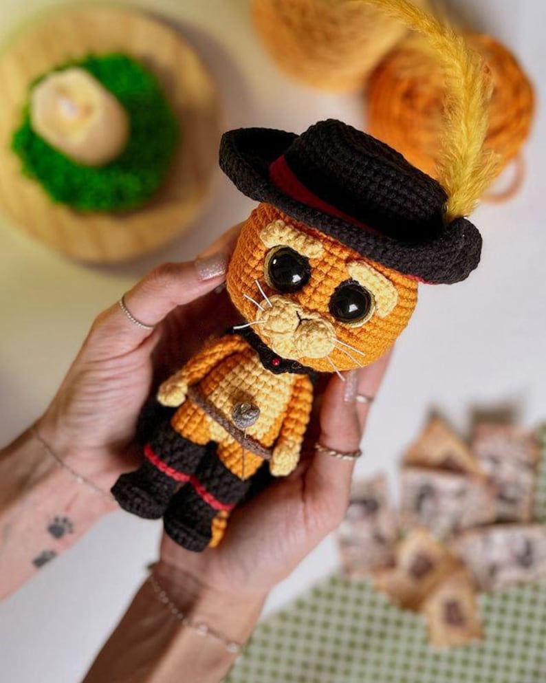 May include: A handmade crochet cat doll, featuring an orange body, black boots, and a black hat with a red band and yellow feather. The doll has large black eyes and white whiskers. The cat is wearing a belt with a buckle.