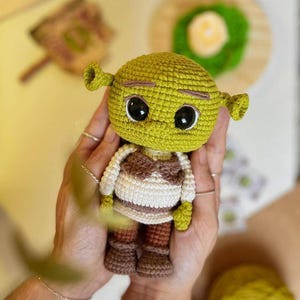 May include: A handmade crochet doll of the character Shrek. The doll has a green head with large black eyes, small ears, and a brown and white outfit. The doll is approximately 5 inches tall.