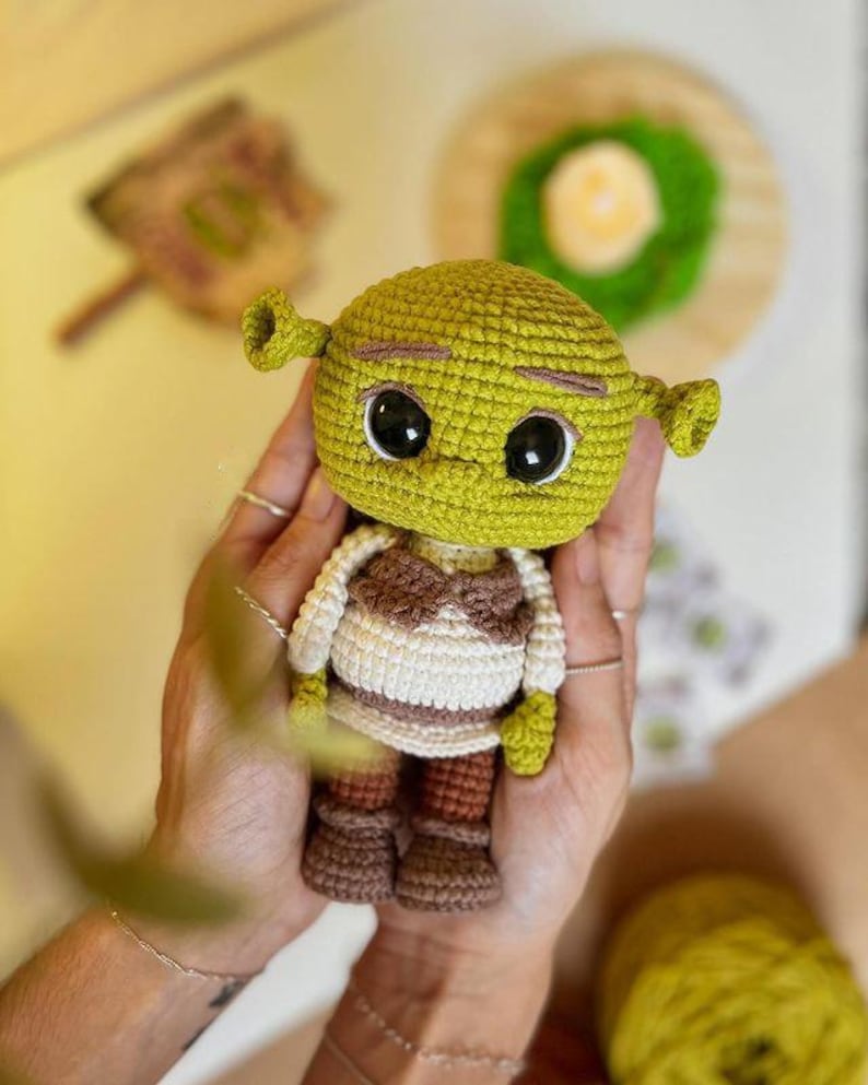 May include: A handmade crochet doll of the character Shrek. The doll has a green head with large black eyes, small ears, and a brown and white outfit. The doll is approximately 5 inches tall.