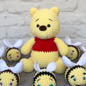 Pode incluir: Um urso de crochê amarelo feito à mão usando uma camisa vermelha, cercado por várias abelhas de crochê. O urso tem um nariz marrom e olhos pretos. As abelhas são amarelas e marrons com asas brancas e olhos verdes. Os brinquedos estão em uma superfície de madeira.