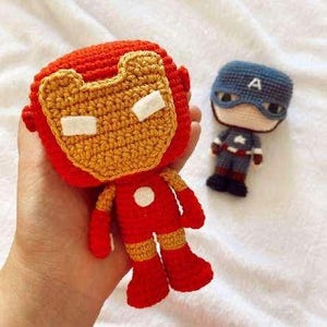 May include: Handmade crochet Iron Man and Captain America dolls. The Iron Man doll is red with a yellow face and white accents. The Captain America doll is blue, gray, and red. Both dolls are on a white surface.