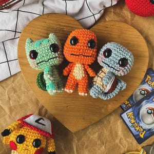 May include: Crocheted Pok&eacute;mon figures of Bulbasaur, Charmander, and Squirtle, along with a Pikachu, are displayed on a wooden heart-shaped board. The figures are in green, orange, and blue, with black eyes. A red and white Pok&eacute;ball and Pok&eacute;mon trading cards are also visible.
