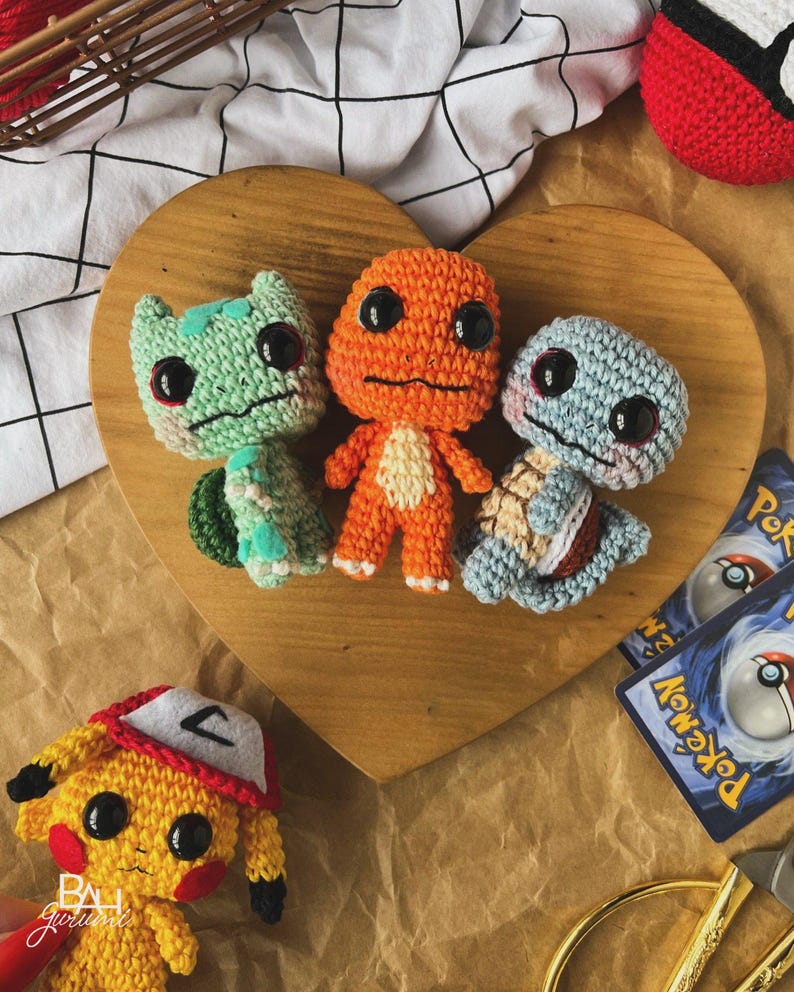 May include: Crocheted Pok&eacute;mon figures of Bulbasaur, Charmander, and Squirtle, along with a Pikachu, are displayed on a wooden heart-shaped board. The figures are in green, orange, and blue, with black eyes. A red and white Pok&eacute;ball and Pok&eacute;mon trading cards are also visible.