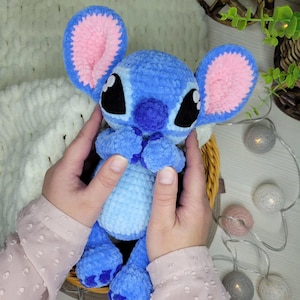 May include: A handmade blue and pink plush toy, resembling a cartoon character, is held in a woven basket. The toy has large ears with pink interiors, black eyes, and a light blue body. The plush is made of soft, textured yarn.