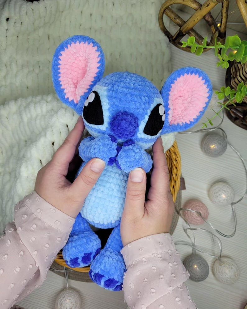 May include: A handmade blue and pink plush toy, resembling a cartoon character, is held in a woven basket. The toy has large ears with pink interiors, black eyes, and a light blue body. The plush is made of soft, textured yarn.
