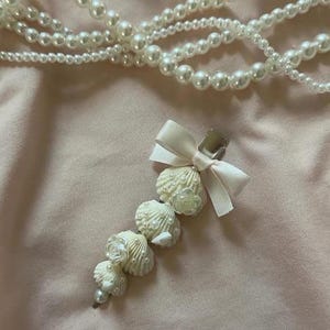 May include: A decorative hair clip featuring a cluster of white seashells and small pearl accents, adorned with a white bow. A strand of white pearls is in the background. The clip is made of metal.