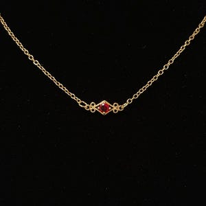 Może przedstawiać: Delikatny złoty naszyjnik z małą, czerwoną zawieszką z kamieniem szlachetnym. Zawieszka ma kształt rombu, a łańcuszek jest w stylu cienkich ogniw. Tło jest jednolicie czarne, podkreślające biżuterię.