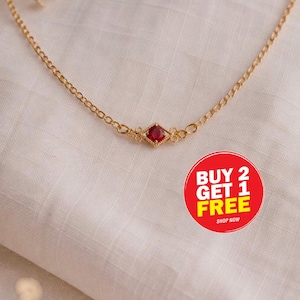 Könnte beinhalten: Eine zarte goldfarbene Halskette mit einem kleinen, quadratischen, roten Edelsteinanhänger. Die Halskette befindet sich auf einem weißen Stoffhintergrund. Ein roter und gelber runder Aufkleber mit dem Text "BUY 2 GET 1 FREE" befindet sich unten rechts.
